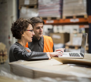 Two team members colloborate in a warehouse while looking at a laptop 